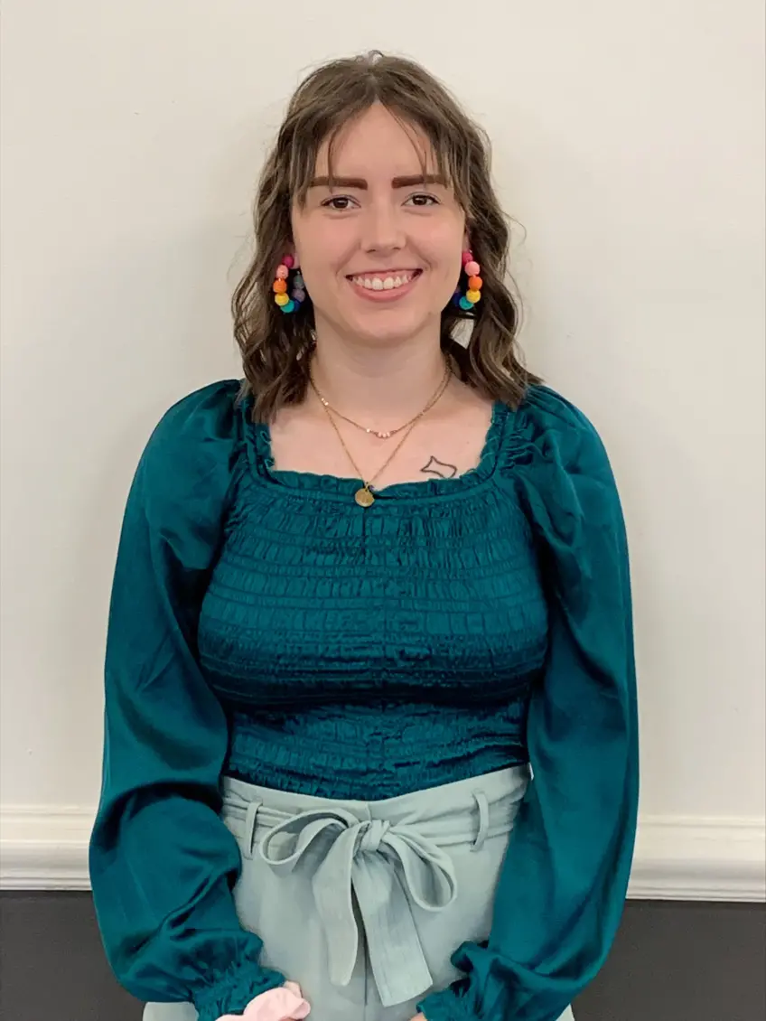 A young woman with long wavy brown hair and brown eyes wearing an aqua blue blouse and colorful hoop earings