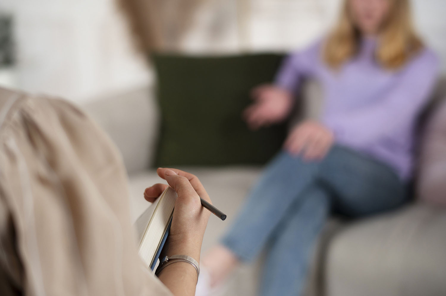 blurred image of the back of a therapist with a pencil and book in her hand and talking sitting across from a female young adult