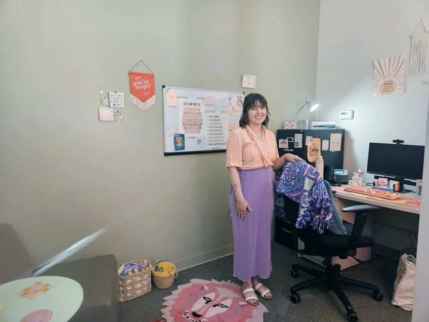 Young female professional wearing lavender pants and a peach shirt and standing in front of her desk in her subtly decorated office.