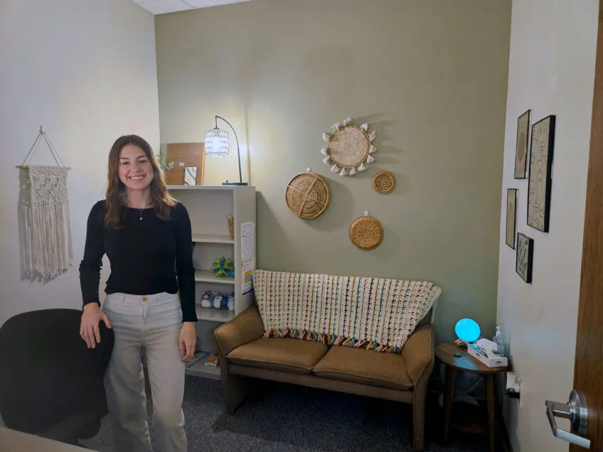 Young adult female wearing a black sweater and tan pants standing in her office with a sofa and blanket and unique straw artwork on the wall