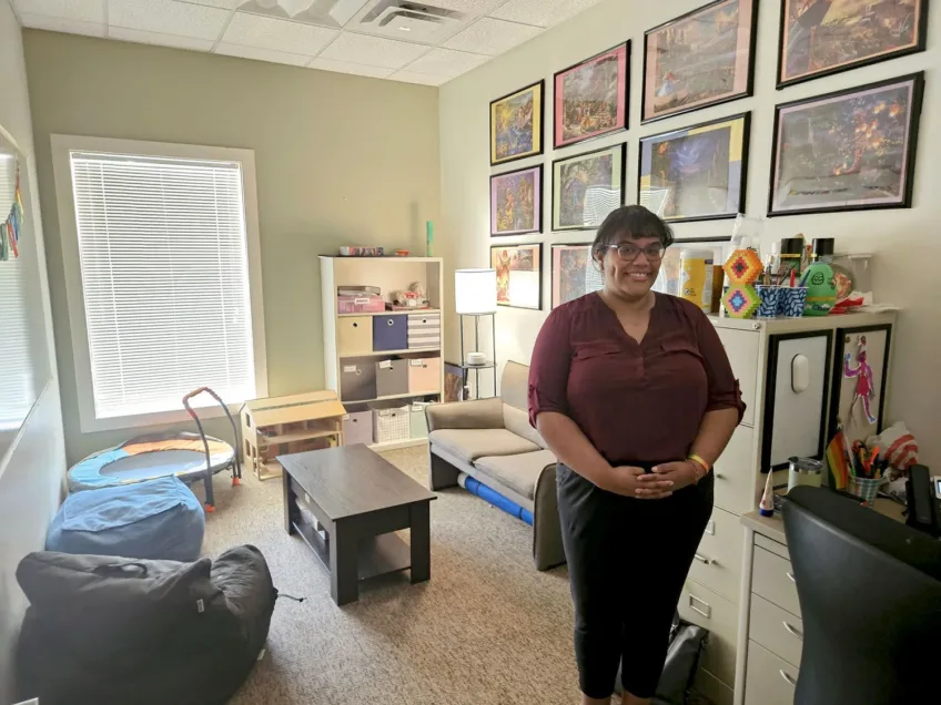 Professional female standing in her comfortably decorated office with colorful pictures on the wall. She is wearing black pants and a maroon blouse.