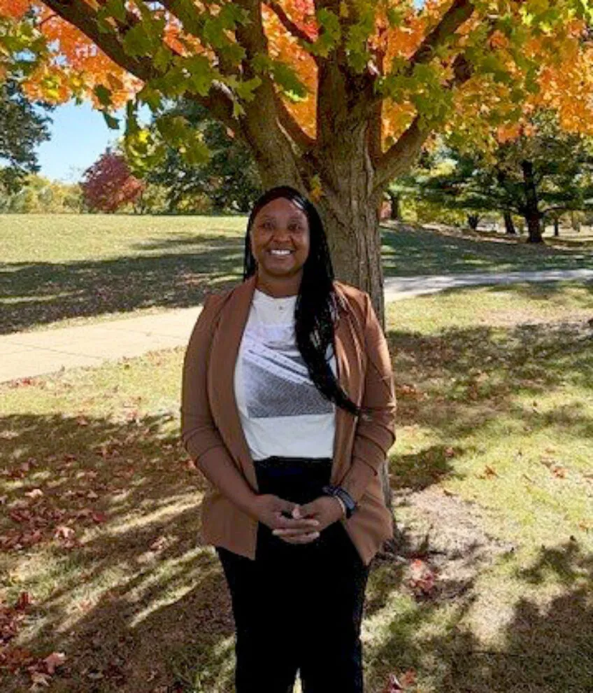 african american young adult in a white tshirt and a cardigan sweater standing in front of a fall tree in a park