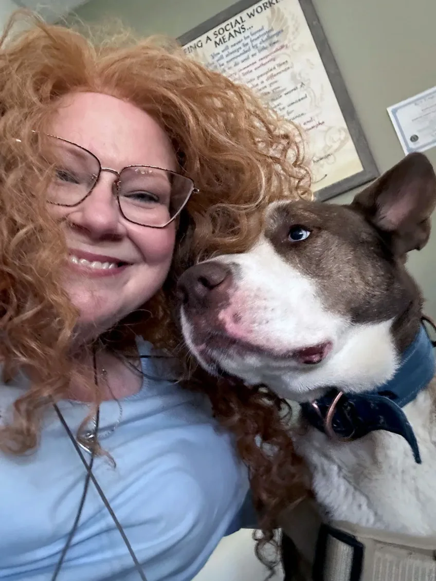 close up selfie of therapist Jennifer who has a light blue shirt on, curly red hair and glasses and she is snuggling up with Lincoln, her Siberian Husky who has a white face but black around the eyes and up through ears