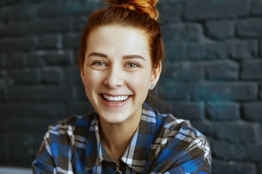 young smiling woman with ginger colored hair in a bun wearing a blue, black and gray flannel shirt and sitting in front of a blue/gray brick wall