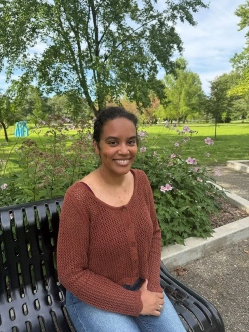 female wearing jeans and a brown sweater, sitting on a bench in a park and smiling at camera